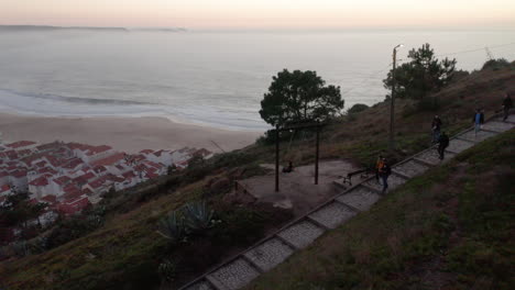 Tourist-Auf-Gehwegweg-Auf-Einem-Hügel-Mit-Blick-Auf-Das-Meer-Und-Die-Altstadt-Bei-Sonnenuntergang-Am-Strand-Von-Nazare,-Portugal