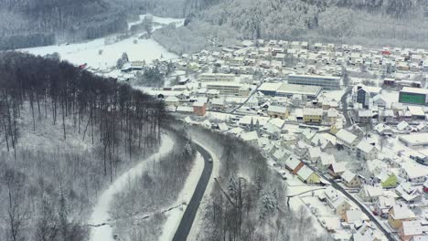 Winter-from-above---drone-camera-is-following-a-car,-which-is-driving-through-a-snowy-landscape-at-a-curve-road-and-a-town-next-to-the-street