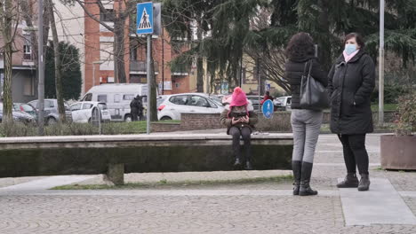 Couple-of-Women-Talking-Outside-in-Town-While-a-Little-Girl-is-Sitting-on-the-Bench-Looking-on-Smartphone