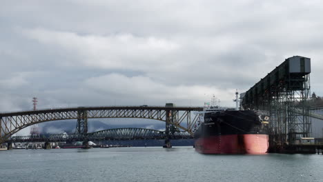 Wide-shot-of-fjord-with-docking-chinese-container-ship-at-pier-and-driving-cars-on-bridge-over-river-during-cloudy-day