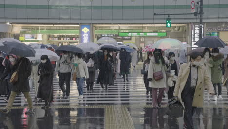 Crowd-Of-People-Wearing-Mask-Crossing-The-Road-With-Umbrella-During-Snowfall-With-Shinjuku-JR-Station-In-The-Background-In-Tokyo,-Japan