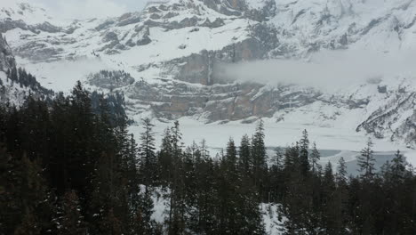 Antena-De-Bosque-Verde-En-Un-Paisaje-Cubierto-De-Nieve-Con-Un-Lago-Congelado-Y-Montañas-En-El-Fondo