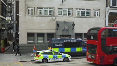 Ein-Polizeiauto-Und-Ein-Lieferwagen-Parken-Vor-Einer-Polizeistation-Im-Zentrum-Von-London,-Während-Ein-Roter-Doppeldeckerbus-Vorbeifährt