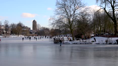 Der-Linke-Schwenk-Zeigt-Die-Malerische-Landschaft-Von-Menschen,-Die-Eislaufen-Und-Sich-Auf-Einem-Zugefrorenen-Kanal-In-Einer-Städtischen-Umgebung-Mit-Kahlen-Winterbäumen-Und-Weißem-Schnee-In-Einer-Kalten,-Gemütlichen-Landschaft-Vergnügen