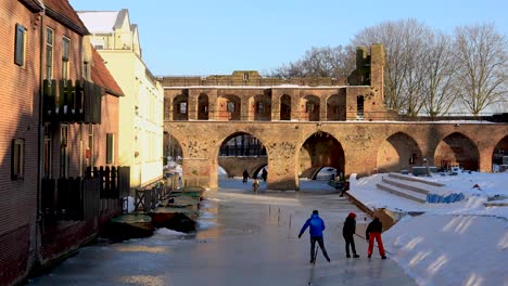 Schneebedeckte-Berkelpoort-Treppen-Und-Jugendliche,-Die-Einen-Eishockeyschläger-Basteln,-Während-Ein-Schlittschuhläufer-Auf-Einem-Zugefrorenen-Fluss-Vorbeifährt,-Der-Unter-Der-Mittelalterlichen-Stadtmauer-Mit-Bootsportal-Vorbeiführt