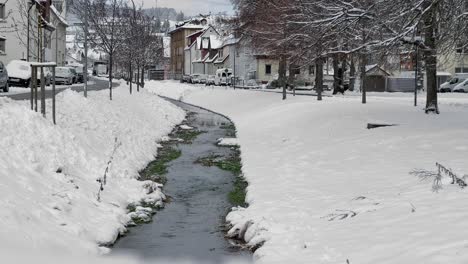 Snowy-river-in-a-idyllic-calm-village-next-to-a-street-with-parking-cars-at-a-bright-winter-day
