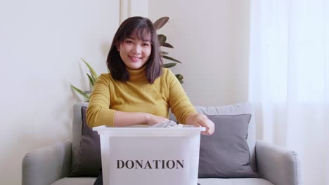 Portrait-of-Asian-young-woman-looking-at-camera-holding-box-with-clothes-for-donation