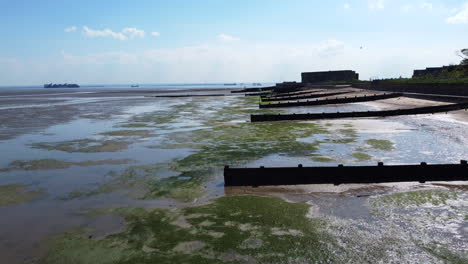 Low-drone-flight-over-estuary-mudflats-and-a-beach-separated-by-old-wooden-groynes-at-low-tide,-with-a-seawall-and-an-abandoned-military-garrison-on-the-right-hand-side,-on-a-clear-and-hot-summer-day