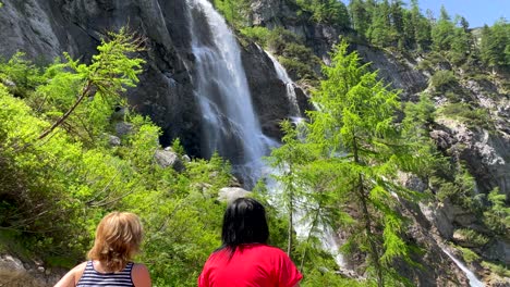 Zwei-Frauen-Bestaunen-Den-Gigantischen-Bergwasserfall,-Der-Im-Sommer-Den-Felsen-Hinunter-Ins-Tal-Stürzt