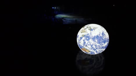Luke-Jarram-Schwebender-Erdbeleuchteter-Planet,-Der-Sich-Nachts-In-Der-Plätschernden-Oberfläche-Des-Seewassers-Spiegelt-Und-Nach-Links-Kreist