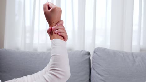 Woman-is-raising-her-hand-to-massage-and-clench-her-sore-wrist-on-the-sofa,-using-her-hands-to-massage-herself-to-relieve-pain-and-improve-symptoms