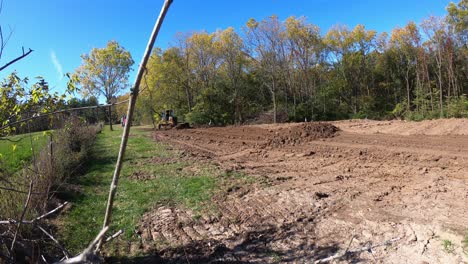 John-Deere-560H-LGP-bulldozer-pushing-black-soil-into-a-small-pile-at-construction-site-with-wooded-area-and-dump-truck-in-background