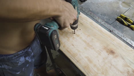 A-shirtless-worker-drilling-hole-into-a-wooden-board-while-making-a-skateboard