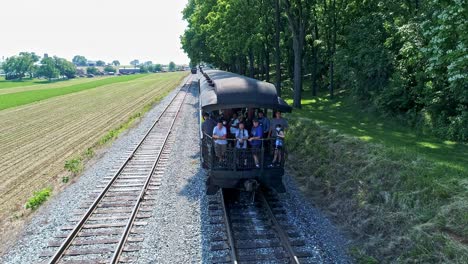 An-Aerial-View-of-a-Steam-Engine-Puffing-Smoke-and-Steam-with-Passenger-Coaches-With-a-Drone-Approaching-the-Back-of-the-last-Coach-with-Travelers-Standing-on-the-Back-of-the-Open-air-Coach-Waving