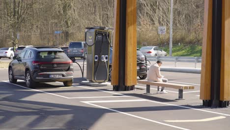 Business-Man-On-Bench-of-Station-Checking-His-Smartphone,-Waiting-Electric-Vehicle-Charging