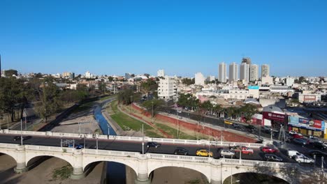 Luftdrohnenüberflug-Puente-Centenario-über-Dem-Fluss-Suquia,-Der-Den-Morgendlichen-Verkehr-Auf-Der-Bogenbrücke-Und-Dem-Boulevard-Mitra-Mit-Der-Innenstadtansicht-Im-Hintergrund-Einfängt