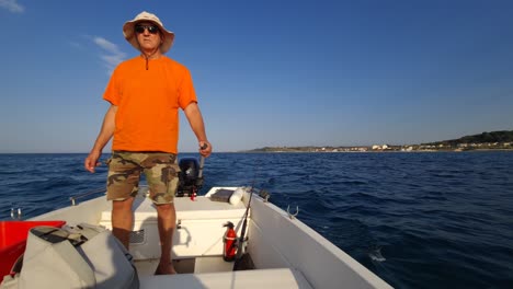 Fisherman-onboard-of-boat-standing-with-sunglasses,-hat,-orange-shirt-and-shorts-fishing-while-moving-and-holding-steering-tiller