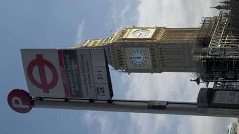 A-vertical-video-of-the-sign-indicating-the-bus-stop-at-Parliament-Square,-the-background-filled-with-the-famous-London-landmark-Big-Ben-and-looking-spectacular-after-recently-being-renovated,-England
