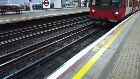 Slow-Motion-shot-surface-metro-train-system-from-London's-station,-with-train-entering-station-as-passengers-wait-on-platform
