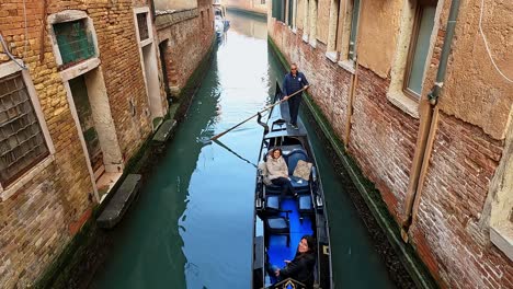High-angle-pov-of-sightseeing-tour-in-gondola-with-seated-tourists-and-gondolier-rowing,-Venice-in-Italy