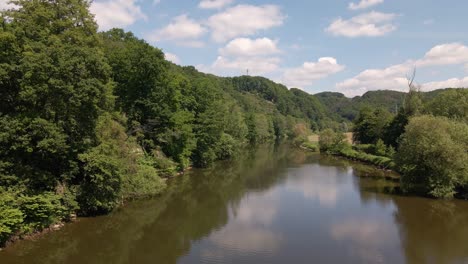 Calm,-brown-river-surrounded-with-lush-and-leafy-riverbanks-underneath-a-blue-sky-with-cumulus-clouds