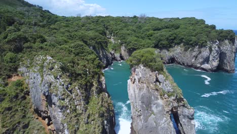 rocky-coastline-of-Asturias-Spain-on-sunny-summer-day-flying-between-large-boulders-in-ocean,-aerial