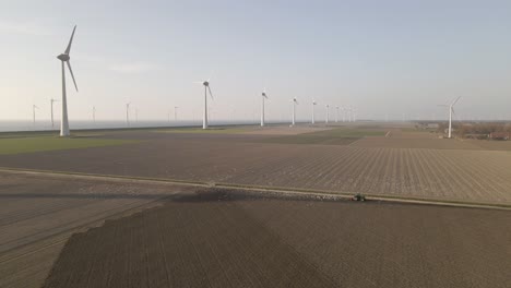 Drone-Shot-of-a-Tractor-Preparing-the-Farm-Fields-on-a-beautiful-day-with-Windmills-in-the-background-and-birds-following