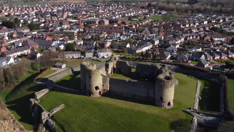 An-aerial-view-of-Rhuddlan-Castle-on-a-sunny-spring-morning,-flying-left-to-right-around-the-castle-with-zoom-out-and-the-town-of-Rhuddlan-in-the-background,-Denbighshire,-Wales,-UK