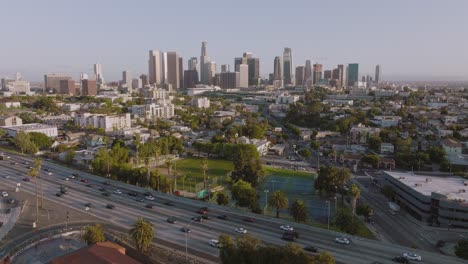 Aerial-Drone-Shot-of-Downtown-Los-Angeles-Skyline,-Busy-Freeway-Below-on-Sunny-Day