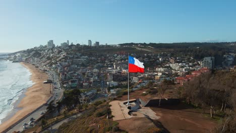 Aerial-reverse-view-republic-of-Chile-flag-flying-on-Reñaca-hilltop-revealing-downtown-city-coastal-buildings