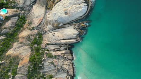 top-down-view-of-large-boulders-on-turquoise-blue-ocean-at-Llandudno-Beach,-aerial
