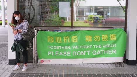 Eine-Frau-Steht-Neben-Einem-Banner,-Das-Die-Öffentlichkeit-Daran-Erinnert,-Versammlungen-Zu-Vermeiden,-Um-Die-Ausbreitung-Des-Ausbruchs-Der-Coronavirus-Variante-In-Hongkong-Einzudämmen