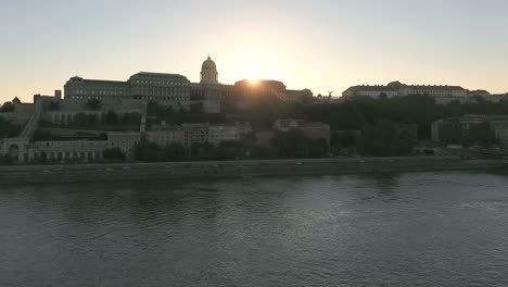 Drone-rising-on-the-Danube,-as-the-setting-sun-appearing-from-behind-the-castle-on-Buda