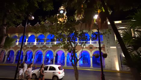 Dolly-shot-to-the-left-through-the-trees-of-the-Municipal-building-at-dusk-with-its-blue-lights-next-to-the-plaza-grande-in-Merida,-Mexico