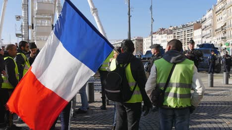 Ein-Demonstrant-Mit-Gelber-Jacke-Und-Französischer-Flagge-Steht-Einer-Reihe-Von-Polizisten-In-Voller-Kampfmontur-Mit-Panzern-Im-Hintergrund-Gegenüber