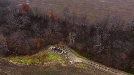 A-drone-flyover-from-high-above-a-pump-jack-working-in-an-old-oilfield-up-against-the-tree-line-adjacent-to-a-harvested-cornfield