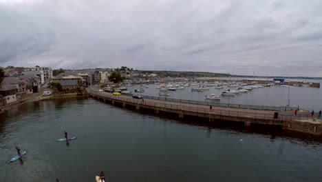 Aerial-footage-of-people-paddle-boarding-in-Penzance-harbour-at-dusk-by-Penzance-Harbour,-dock,-boats-and-yachts-in-the-beautiful-picturesque-area-of-Cornwall,-popular-with-Holiday-makers