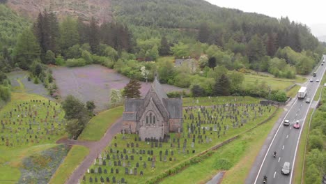 Traffic-on-road-near-St-John's-Episcopal-Church,-Ballachulish-in-the-Scottish-Highlands-surrounded-with-graves