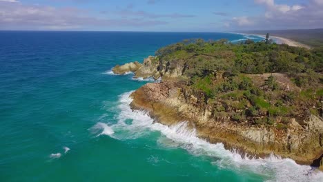 Aerial-view-of-a-rocky-cliff-with-turquoise-blue-water,-vegetation,-waves-splashing