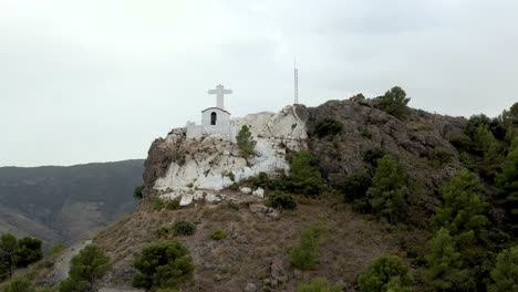 Tourists-on-rocky-mountain-top-with-tiny-white-church-in-Spain,-low-parallax-drone