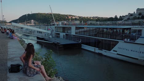 Vista-Del-Danubio-Desde-La-Orilla-De-Raqpart,-Lado-De-Plagas,-Niña-Sentada-Frente-Al-Crucero-Por-El-Río-Crystal-Mahler