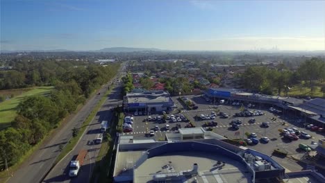 Aerial-drone-shot-moving-past-a-local-shopping-centre-towards-a-busy-arterial-road,-with-Mount-Coot-Tha-and-downtown-Brisbane-in-the-distance