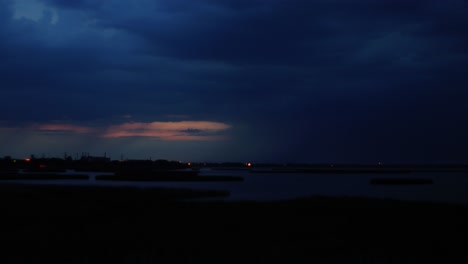 Time-lapse-of-dramatic-dark-thunderstorm-clouds-with-lightning-strikes-rolling-over-the-city-and-lake-Liepaja-after-the-sunset,-wide-shot
