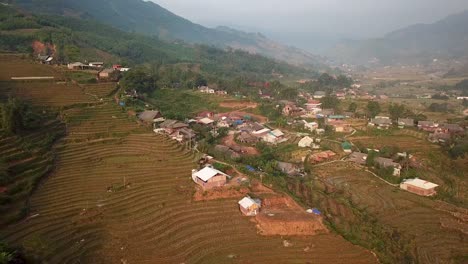 Aerial-view,-pull-back-revealing-rural-neighborhood-surrounded-by-amazing-rice-terraced-green-misty-mountainside-in-Sapa,-Vietnam-at-dusk-as-sun-sets