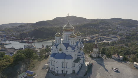 Toma-Aérea-De-Descenso-Lento-De-Una-Iglesia-Ortodoxa-Con-Techo-Azul-Y-Cúpulas-Doradas,-Ubicada-En-La-Cima-De-La-Colina-Con-La-Revelación-Del-Puerto-Y-El-Edificio-De-La-Ciudad-En-El-Fondo,-En-Un-Día-Brillante,-Claro-Y-Soleado