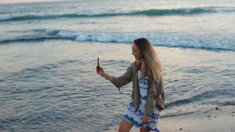 Hübsche-Junge-Blonde-Frau,-Die-Bei-Sonnenuntergang-Am-Strand-Steht,-Selfie-Fotos-Und--Videos-Macht,-Tanzt-Und-Lächelt