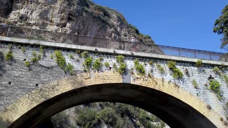 Arch-bridge-with-man-with-white-hat-on-the-railing-while-bus-with-tourists-stops,-Aerial-orbit-track-shot