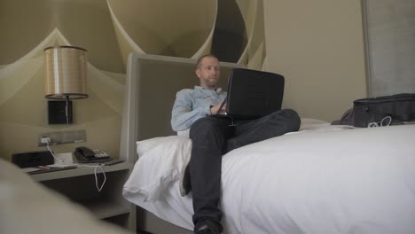 Ein-Weißer-Mann-Mittleren-Alters-Sitzt-Oder-Liegt-Zusammengesunken-Im-Bett-Seines-Modernen-Hotelzimmers-Und-Arbeitet-An-Seinem-Laptop