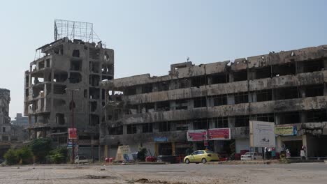 ángulo-Bajo-De-Disparo-Constante,-Autos-Que-Pasan-Por-Un-Enorme-Edificio-Abandonado-Y-Destrozado-En-La-Ciudad-De-Homs