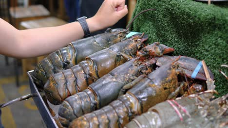 Man-using-arm-to-compare-sizes-fresh-lobsters-on-street-food-market-stall,-Thailand